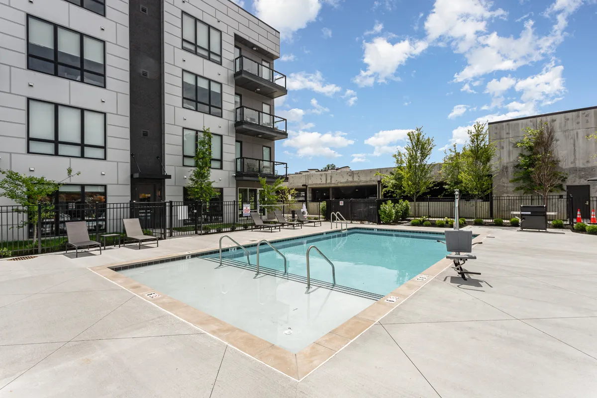 A sparkling resort-style swimming pool is the focal point, with balconies of the building and landscaped greenery surrounding the area. The space is filled with sunlight, suggesting a relaxing, inviting atmosphere ideal for families and groups to enjoy together.