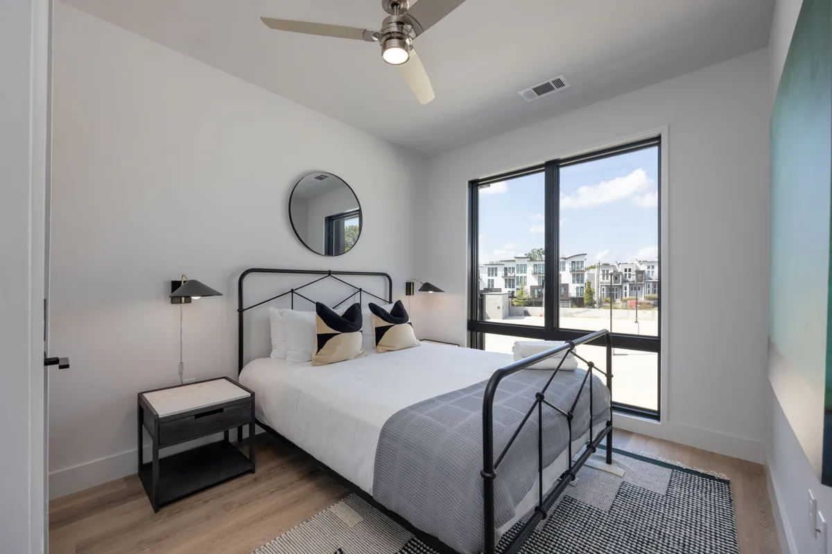 A cozy second bedroom with a plush bed adorned with decorative pillows, a large window letting in daylight, a circular mirror, and a well-placed nightstand and lamp. The atmosphere is inviting and perfect for relaxation or reading.