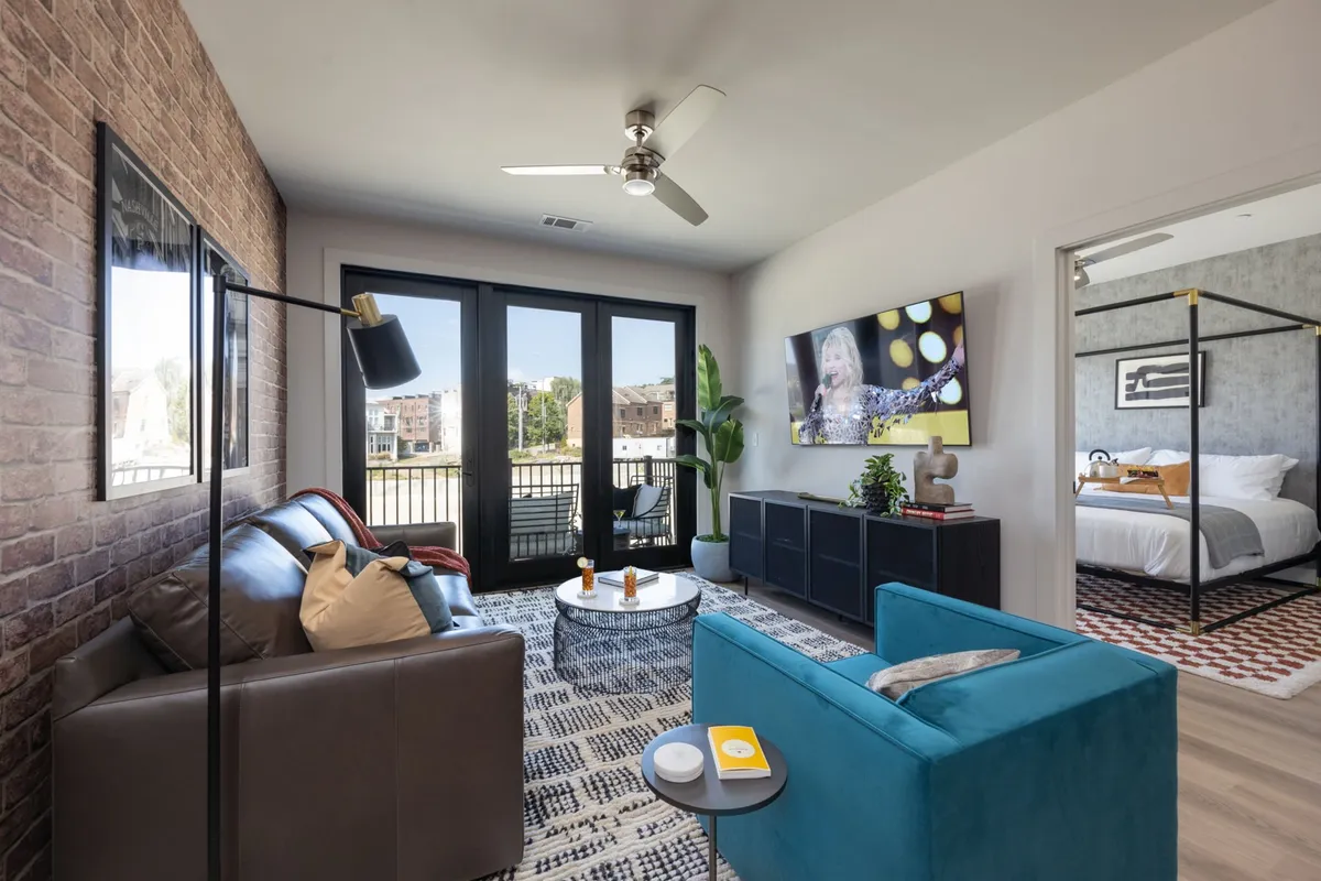 The image shows a stylish, contemporary living room featuring a rich brown leather sofa, a vibrant teal accent chair, a large flat-screen TV, and glass sliding doors that fill the space with natural light. A decorative plant adds a touch of greenery, creating a cozy and inviting atmosphere ideal for relaxing, socializing, or enjoying a quiet movie night.
