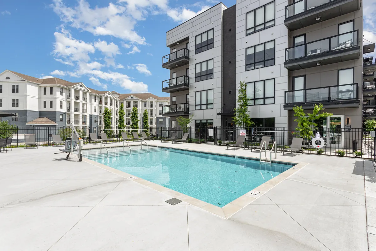 An outdoor pool scene with balconies of the apartments overlooking the water, green trees providing shade, and lounge chairs arranged for comfort. Residents are shown enjoying the pool, socializing, and basking in a refreshing, resort-like ambiance.