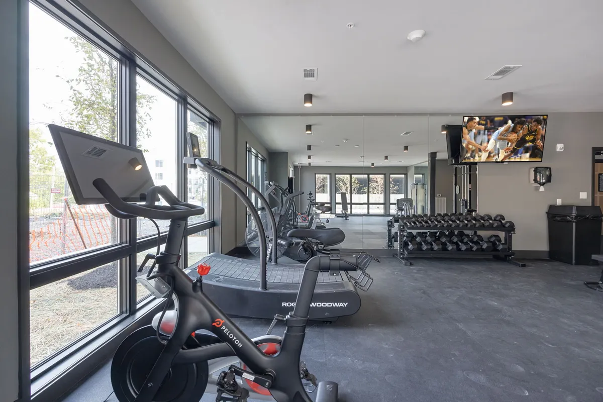 This interior shot showcases a modern fitness center equipped with a Peloton bike, treadmill, dumbbell rack, and large windows flooding the space with natural light. The well-organized layout and sleek equipment promote an energetic and communal workout environment.
