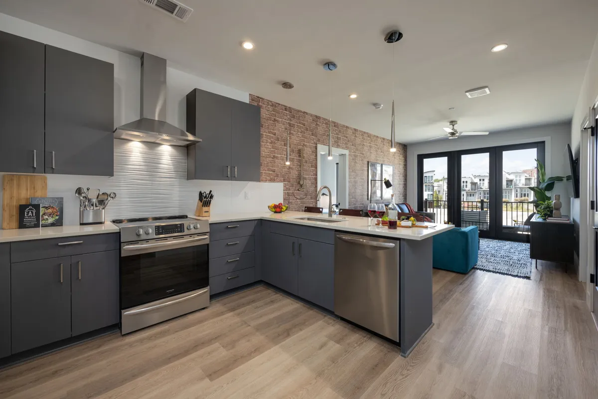 A warm, contemporary kitchen with a prominent kitchen island, stainless steel appliances, exposed brick wall, and large windows. The scene suggests an inviting atmosphere for both cooking and entertaining, with plenty of space for meal preparation and social interaction.