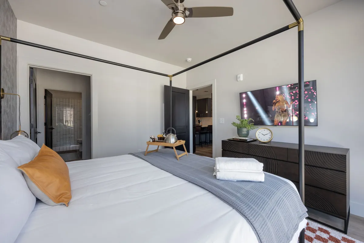 A serene bedroom featuring a canopy bed with crisp white linens, a nightstand with a tray, and a wall-mounted television. The setup exudes a peaceful atmosphere and invites guests to unwind, with plenty of natural light filtering through large windows.