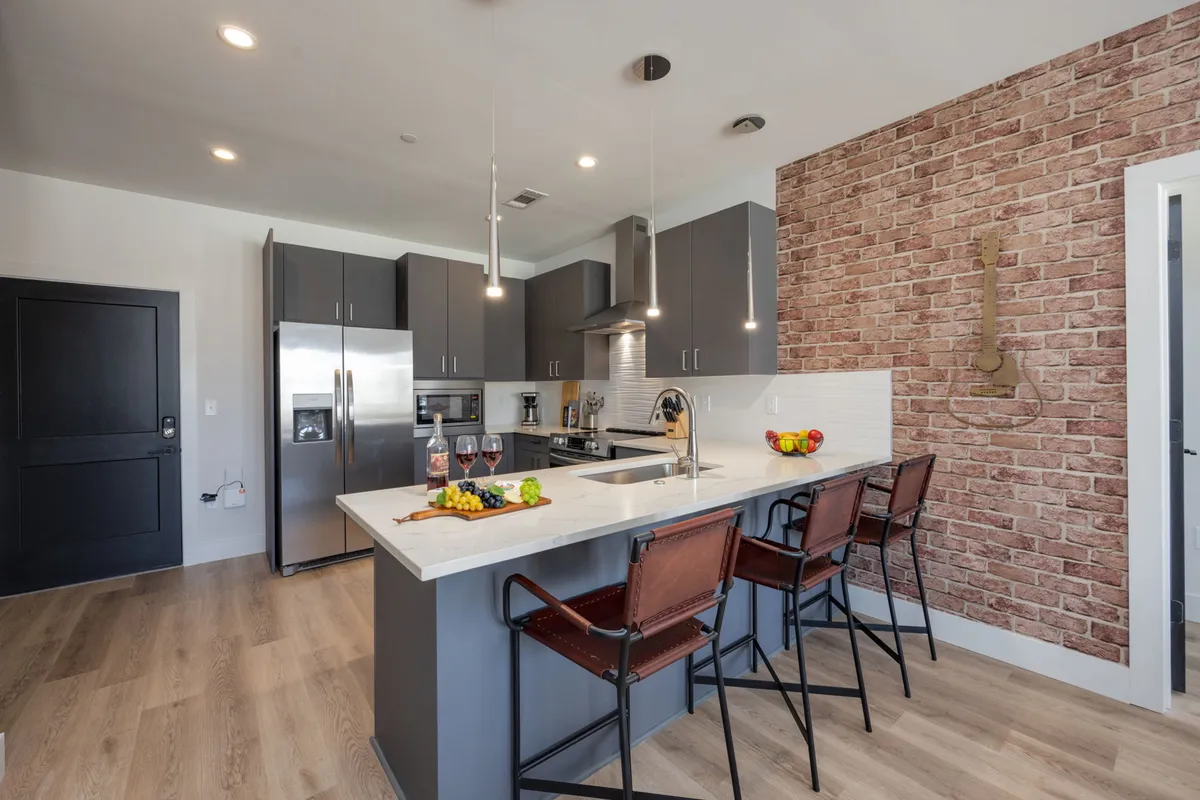 A contemporary kitchen and dining area with a stylish kitchen island, bar stools, exposed brick accent wall, and countertops adorned with fruits and drinks. The space is ideal for both meal preparation and casual dining, seamlessly blending into the open living area.
